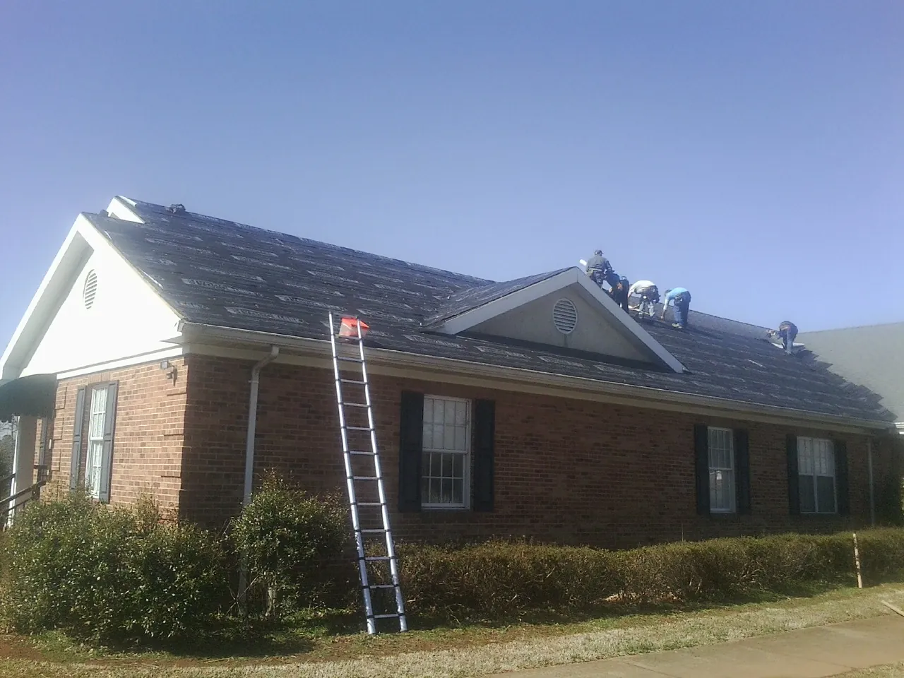 Crew tearing shingles off a brick ranch roof with a ladder set — Emergency Roof Repair work by our crew in Sagamore