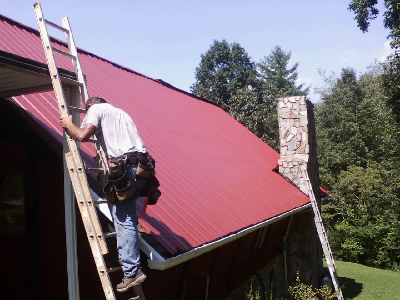 Roofer on a ladder beside a red metal roof and stone chimney — Soffit & Fascia Repair by Roofing Brothers in Sagamore